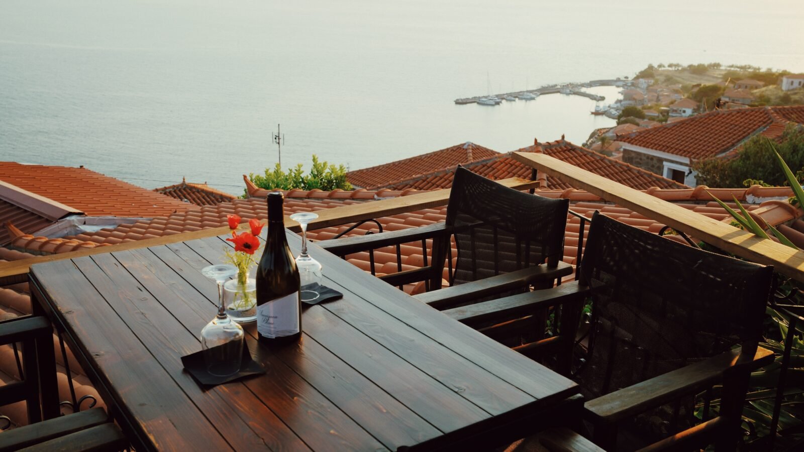 Rooftop terrace in Molyvos with a bottle of wine, two glasses, and a sunset view over the harbor and red-tiled roofs
